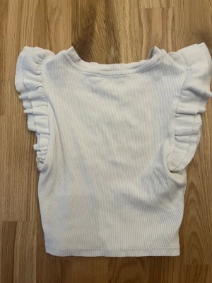 Vit topp med volang från Zara - Supersöt vit ribbad topp från Zara med volangdetaljer längs axlarna. Perfekt att använda nu till sommaren 
