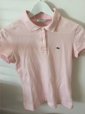 Ljusrosa pikétröja från Lacoste - Säljer en ljusrosa pikétröja från Lacoste med klassisk krage och två knappar framtill. Tröjan har korta ärmar och den ikoniska gröna krokodilloggan på bröstet. Använd fåtal gånger och har inga fläckar på sig. Den är ganska liten för att vara en 36 så jag skulle säga att den är emellan strl 34-36. Pris kan diskuteras😊