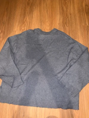 Blå stickad tröja i storlek M - En enkel och stilren grå stickad tröja med rund hals och långa ärmar. Tröjan har ribbstickade muddar vid ärmslut och nederkant. Perfekt för lager på lager och passar till det mesta i garderoben.
