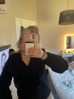 Svart blus med volangdetaljer - Snygg svart blus med v-ringning och volangdetaljer längs axlarna. Blusen har långa ärmar med resår vid ärmslut och är tillverkad i ett lätt, något transparent tyg som ger en luftig känsla. Perfekt att styla med både jeans och kjol.