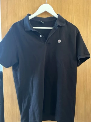 Svart pikétröja med liten logga - Klassisk svart pikétröja i bomull med korta ärmar och krage. Tröjan har en liten rund logga på bröstet och knäppning vid halsen. Perfekt till jeans eller shorts för en enkel och stilren look.