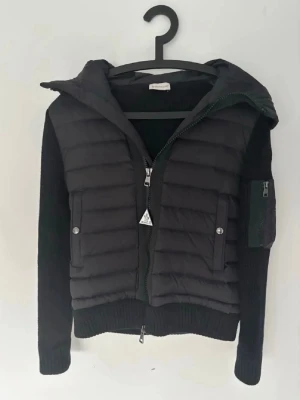  Moncler Cardigan - Snygg svart cardigan från Moncler med stickade ärmar och stor vadderad huva. Skicket är 10/10 då den knappt är använd. Västen har två fickor med tryckknappar framtill, dragkedja och en extra ficka med dragkedja på ärmen. Perfekt för lager-på-lager. Vid snabb affär kan jag sänka priset. Sitter perfekt som S då de står l