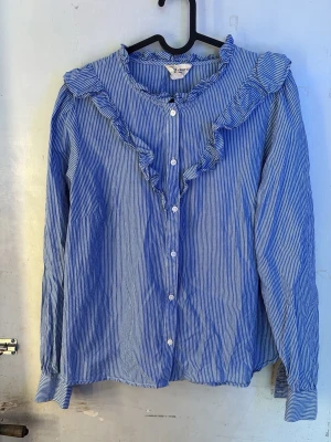 Blårandig blus med volangkrage - Söt blus/skjorta  med vita smala ränder. Blusen har långa ärmar, knäppning framtill och en stor volangkrage som ger en trendig look. Tillverkad i ett lätt bomullstyg som känns skönt mot huden. Perfekt för dig som gillar romantiska detaljer. Super fin nu till vår! Den är ostruken nu men när man stryker den blir den så fin!