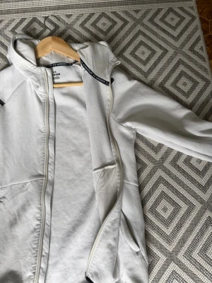 Vit hoodie från Under Armour - Snygg vit hoodie från Under Armour med dragkedja framtill och huva. Hoodien har svarta detaljer, två fickor på sidorna och en liten ficka med dragkedja på ärmen. Tillverkad i mjukt och bekvämt material, perfekt för en sportig och avslappnad stil.