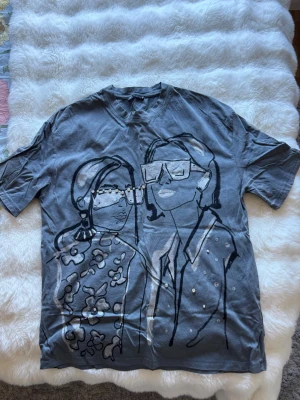 Grå t-shirt med konstnärligt tryck - Säljer en grå t-shirt med ett stort konstnärligt tryck av två personer på framsidan. T-shirten har korta ärmar och detaljer med pärlor på glasögonen och kläderna i motivet. Perfekt för dig som gillar unika plagg med cool design.