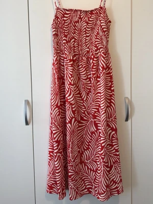 Rödvit långklänning med mönster - Snygg långklänning med röda och vita bladliknande mönster. Klänningen har smala axelband och är ärmlös, med en smockad överdel som ger en bekväm passform. Tillverkad i mjuk bomull, perfekt för varma sommardagar.