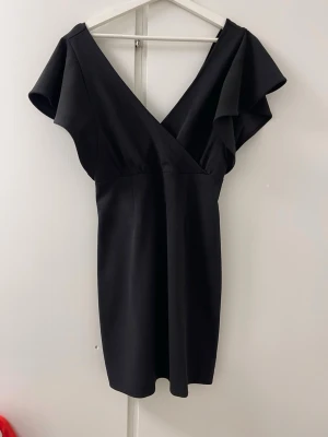 Svart omlottklänning med volangärm - Stilren svart kortklänning med omlott-ringning och volangärmar. Klänningen har en figurnära passform och är tillverkad i ett mjukt, slätt material. Perfekt för dig som gillar en enkel men elegant look.