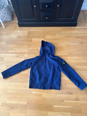 Blå vindjacka från Stone Island - Snygg blå vindjacka från Stone Island med huva och klassisk logotyp-patch på vänster ärm. Jackan har dragkedja framtill och är tillverkad i ett lätt, vindtätt material som passar perfekt för blåsiga dagar. Enkel och stilren design som funkar till det mesta.