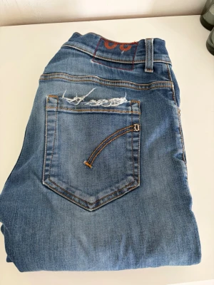 Blå slitna jeans från dondup - Säljer ett par blå jeans från dondup Jeans med slitningar och hål på benen för en trendig, avslappnad look. Jeansen har klassisk femficksmodell, kontrastsömmar och en patch bak i midjan. Perfekta för dig som gillar streetstyle och en casual vibe.