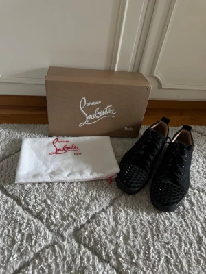 Svarta sneakers - Svarta loubs med klassiska röda sulor och coola nitar på tån. Skorna har snörning och är tillverkade i skinn med en lyxig känsla. Perfekta för dig som vill sticka ut med en exklusiv och edgy stil.