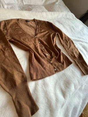 Brun kofta med v-ringning och knappar - Snygg brun kofta med v-ringning och långa ärmar. Koftan har ribbade muddar och stängs med fem ljusa knappar framtill. Perfekt att slänga över en t-shirt eller linne för en chill look.