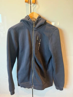 Marinblå hoodie från Sail Racing - Snygg marinblå hoodie från Sail Racing med dragkedja framtill och en bröstficka med logga. Tröjan har huva med dragsko och ribbade muddar vid ärmslut och nederkant. Perfekt för en avslappnad och sportig stil. Materialet känns mjukt och bekvämt.
