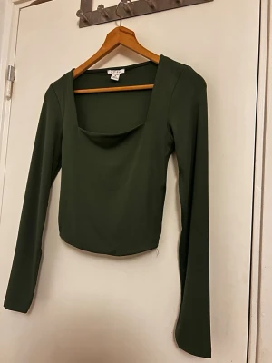 Mörkgrön långärmad topp med fyrkantig ringning - Snygg mörkgrön långärmad topp med fyrkantig ringning och croppad passform. Perfekt att styla med höga jeans eller kjol. Tillverkad i mjukt stretchigt material för en bekväm känsla. Aldrig använt då jag inte tar på mig croppat
