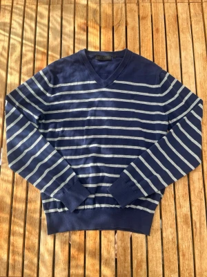 Marinblå randig stickad tröja - Snygg marinblå stickad tröja med vita horisontella ränder och V-ringning. Tröjan har långa ärmar och ribbade muddar vid ärmslut och nederkant. Perfekt för dig som gillar klassisk och stilren look.