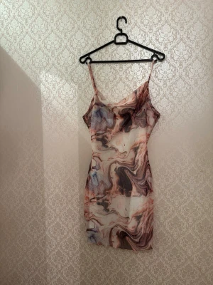 Mönstrad kortklänning med tunna band - Säljer en kort klänning med tunna axelband och ett unikt marmorerat mönster i beige, brun, svart och blå toner. Klänningen har en figurnära passform och är perfekt för dig som gillar trendiga och färgstarka plagg. Materialet känns lätt och svalt.