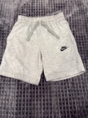 Grå shorts från Nike med snörning - Snygga grå shorts från Nike med elastisk midja och snörning framtill. Klassisk Nike-logga broderad på vänster ben. Tillverkade i mjuk bomullsmix för skön känsla. Perfekta för avslappnade dagar eller träning.