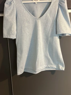Ljusblå glittrig topp med puffärm - Supersöt ljusblå topp med glittrigt tyg och puffiga korta ärmar. Djup v-ringning framtill ger en trendig look. Perfekt för dig som vill sticka ut lite extra. 