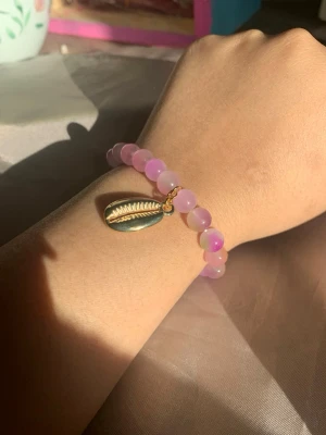 Rosa pärlarmband med snäckberlock - Snyggt armband med ljusrosa pärlor och en guldfärgad snäckberlock. Pärlorna är runda och skimrande, vilket ger ett fräscht och trendigt intryck. Perfekt accessoar för dig som gillar strandvibbar och unika detaljer.