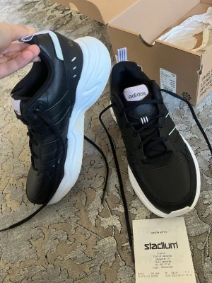 Svarta Adidas sneakers med vit sula - Stilrena svarta sneakers från Adidas med chunky vit sula och klassiska ränder på sidan. Ovandelen är i syntetmaterial och textil, med snörning och dragflik bak. Perfekta för dig som gillar sportig och modern look. Kvitto ingår 