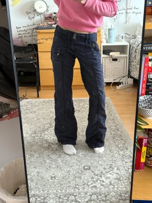 Mörkblå cargobyxor med fickor - Säljer ett par mörkblå cargobyxor med raka ben och flera praktiska fickor på sidorna. Byxorna har dubbla knappar i midjan och detaljerade sömmar för en cool look. Perfekta för dig som gillar streetwear och funktionella plagg. Dom har några få defekter - se bilder 