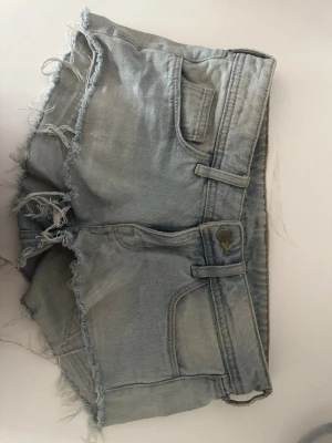 Ljusblå jeansshorts med fransar - Säljer ett par ljusblå jeansshorts från H&M med råa, fransiga kanter och klassisk femficksdesign. Shortsen har låg midja och en avslappnad passform. Perfekta för varma dagar och en cool, avslappnad stil.