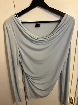 Ljusblå långärmad topp Gina Tricot - Säljer en ljusblå långärmad topp från Gina Tricot med draperad halsringning och figurnära passform. Toppen har en mjuk och stretchig känsla.
