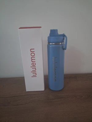 Blå vattenflaska från Lululemon - Snygg blå vattenflaska från Lululemon i rostfritt stål med praktiskt skruvlock och handtag. Flaskan har en stilren design med Lululemon-loggan ingraverad på sidan. Perfekt att ta med till gymmet, skolan eller på utflykten.