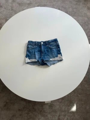 Blå jeansshorts med spetsdetaljer - Snygga blå jeansshorts med rå kant och vita spetsdetaljer vid bensluten. Klassisk femficksmodell med bälteshällor och knappstängning. Shortsen är i storlek 152 och är i bra skick❣️. Perfekta för varma dagar och ger en avslappnad men söt look.