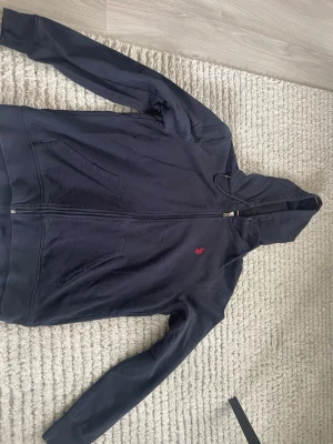 Mörkblå hoodie från Ralph Lauren - Mörkblå hoodie med dragkedja från Ralph Lauren. Klassisk design med huva och två fickor framtill. Liten röd broderad logga på bröstet. Tillverkad i mjukt bomullsmaterial som känns skönt mot huden. Perfekt för en avslappnad och stilren look. Passar väldigt bra för vår väder också. PRISET KAN DISKUTERAS!