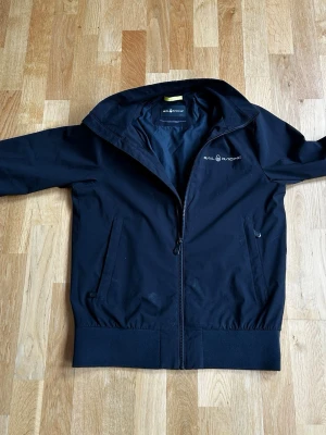 Svart vindjacka från Sail Racing - Stilren svart vindjacka från Sail Racing med dragkedja framtill, två sidofickor och diskret logga på bröstet. Jackan har hög krage och breda muddar vid ärmslut och nederkant. Perfekt för blåsiga dagar och enkel att matcha med olika outfits.