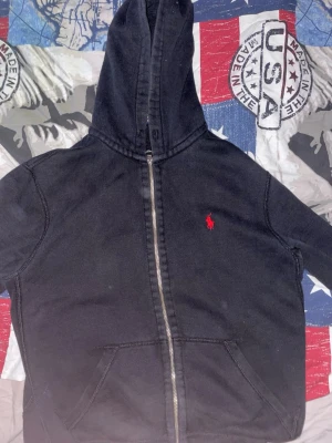 Svart hoodie från Polo Ralph Lauren - Svart hoodie med dragkedja från Polo Ralph Lauren. Klassisk design med röd broderad logga på bröstet, huva och kängurufickor framtill. Tillverkad i mjukt material som känns skönt mot huden. Perfekt för en avslappnad och stilren look.