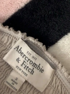 Beige kort klänning Abercrombie - Säljer en beige kort klänning från Abercrombie & Fitch i storlek S. Klänningen har tunna axelband, fickor framtill och volangkant nedtill. Materialet har en lätt glansig finish och smock upptill för en bekväm passform.