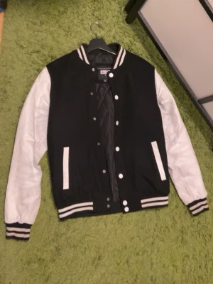 Svart och vit bomberjacka - Snygg svart och vit bomberjacka från H&M med klassisk collegestil. Jackan har vita ärmar, svart kropp och ribbade muddar med vita ränder. Knäppning med vita tryckknappar och två fickor fram. Perfekt för en sportig och trendig look.