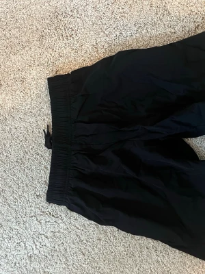 Svarta shorts med snörning - Svarta shorts med resårmidja och snörning. På ena benet står texten 'YOU CAN JOIN US' tryckt i svart. Shortsen har en loose passform och är perfekta för träning eller chill. Tillverkade i ett lätt syntetmaterial.