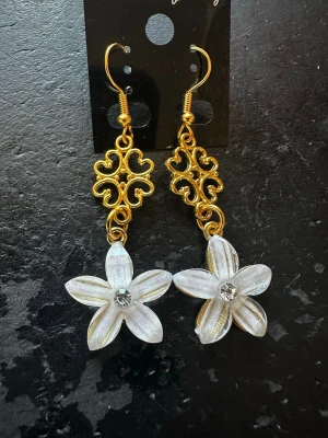 Blommiga örhängen med pärldetaljer - Unika örhängen med guldfärgade detaljer och vita blommor i plast. Blommorna har en glittrande sten i mitten som ger extra bling. Hängena har en elegant, snirklig design upptill och krokar för enkel användning. Perfekta för dig som vill sticka ut med en söt och trendig accessoar.