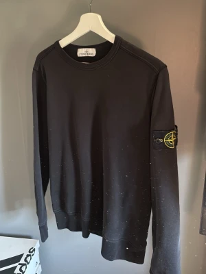 Svart sweatshirt från Stone Island - Svart sweatshirt från Stone Island med klassisk rund halsringning och ikonisk logopatch på vänster ärm. Tröjan har en avslappnad passform och ribbade muddar vid ärmslut och nederkant. Perfekt för en streetwear-look.