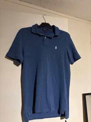 Blå pikétröja från Polo Ralph Lauren - Snygg blå pikétröja från Polo Ralph Lauren med klassisk krage, två knappar och vit broderad logga på bröstet. Tillverkad i mjuk bomull och har korta ärmar för en avslappnad men stilren vibe.