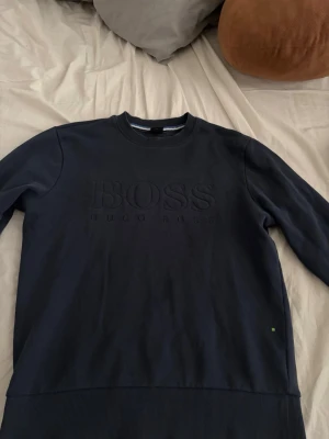 Mörkblå sweatshirt från Hugo Boss - Mörkblå sweatshirt från Hugo Boss med diskret broderad logga på bröstet. Tröjan har rund halsringning, långa ärmar och ribbade muddar vid ärmslut och nederkant. Perfekt för en stilren och avslappnad look.