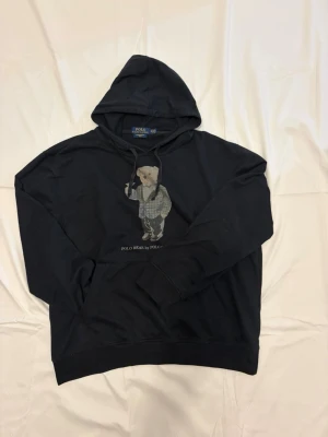 Svart hoodie Polo Bear från Polo Ralph Lauren - Svart hoodie från Polo Ralph Lauren med ikoniska Polo Bear-motivet på bröstet. Klassisk huva med dragsko och ribbade muddar. Materialet är mjukt och bekvämt, perfekt för en avslappnad stil. Snyggt tryck med björn i kostym på framsidan. Har tvättats för varmt så skulle uppskatta den som en M nu.