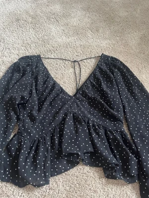 Svart blus med vita prickar och volang - Svart blus med vita prickar, v-ringning och knytband i nacken. Blusen har långa ärmar och volang nertill, vilket ger en luftig och trendig look. Perfekt för dig som gillar en söt och stilren stil.
