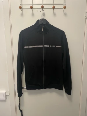 Svart zip-hoodie från Hugo boss - Svart hoodie från BOSS med hel dragkedja framtill och diskret logga på bröstet. Har en grå rand över bröstet och två fickor framtill. Klassisk design med hög krage och ribbade muddar. Perfekt för en stilren och avslappnad look.