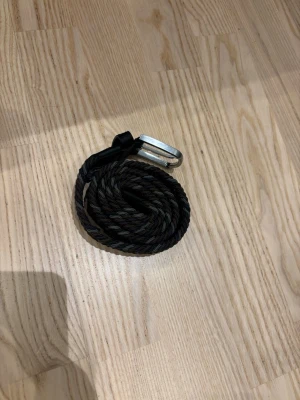 Svart flätat bälte med silverdetalj - Snyggt svart flätat bälte med silverfärgat spänne och detaljer i svart skinn. Perfekt accessoar för att lyfta din outfit och funkar till både jeans och byxor. Enkel att justera tack vare det flexibla materialet.