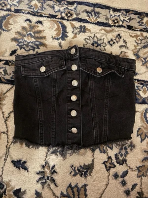Svart jeans topp från FB Sister - Cool svart bustier från FB Sister i denim med knappar framtill och rå fransad nederkant. Toppen har två bröstfickor med knapp och markerade sömmar som ger en snygg siluett. Perfekt att styla med höga jeans eller kjol.