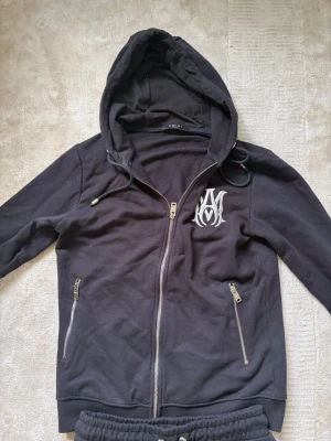 Svart tracksuit set från Amiri, Oanvänd.  - Svart hoodie och byxor från Amiri med hel dragkedja, huva med dragsko och två fickor med dragkedja. Stort vitt Amiri-logotyptryck på bröstet. Tillverkad i mjukt bomullsmaterial, perfekt för en avslappnad streetwear-look. Produkten är oanvänd och har bara legat i garderoben, storlek Medium. 