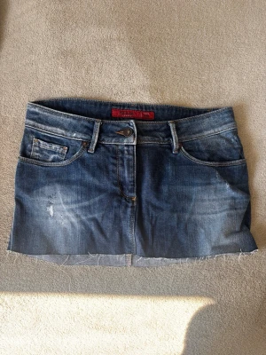 Jeanskjol  - Cool jeanskjol från Diesel i klassisk blå denim med rå, avklippt kant. Kjolen har fem fickor, bälteshällor och slitna detaljer för en avslappnad look. Perfekt för dig som gillar streetstyle och denim.