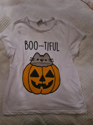 Vit Pusheen t-shirt med pumpa - Söt vit t-shirt från Pusheen med tryck av en katt och en stor orange pumpa med texten 'BOO-TIFUL' på bröstet. Perfekt för dig som älskar gulliga motiv och vill ha en rolig t-shirt till hösten eller halloween. Tillverkad i mjuk bomull och har korta ärmar.