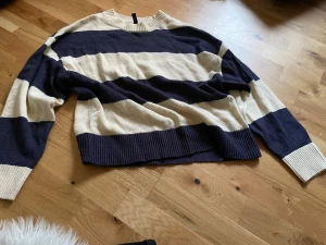 Randig stickad tröja i blått och beige - Säljer en oversized stickad tröja med breda horisontella ränder i mörkblått och beige. Tröjan har rund halsringning, långa ärmar och ribbstickade muddar vid ärmslut och nederkant. Perfekt för en avslappnad och trendig look.