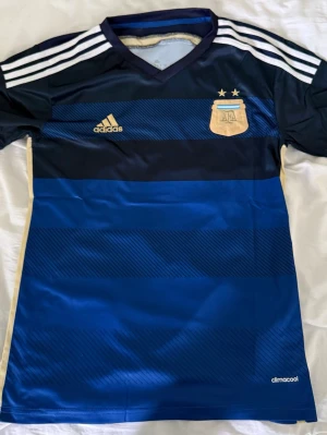 Argentina Adidas fotbollströja blå - Snygg blå fotbollströja från Adidas med Argentinas landslagsemblem och två stjärnor på bröstet. Tröjan har mörkblå och ljusblå ränder, vita axelränder och guldiga detaljer. Tillverkad i lätt och ventilerande climacool-material.