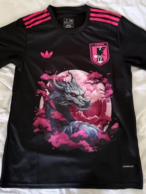 Japan Adidas svart tröja med drake - Svart sporttröja från Adidas med rosa detaljer och Japan JFA-logga. Framsidan har en stor grafisk drake, rosa körsbärsblommor och måne. Tre rosa ränder på axlarna och Aeroready-material för bra andningsförmåga. Perfekt för dig som gillar unika sportkläder.