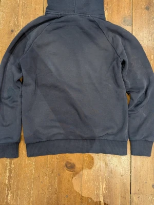 Mörkblå hoodie från Peak Performance - Snygg mörkblå hoodie från Peak Performance med vit logga och text på bröstet. Klassisk känguruficka framtill, ribbade muddar och justerbar huva med snören. Perfekt för en avslappnad och sportig stil.